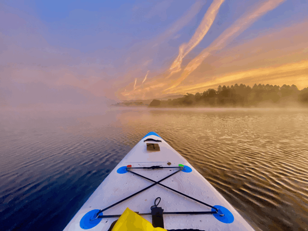 Front of a white paddle board on a calm misty lake at sunset with woodland lining the distant lake shore