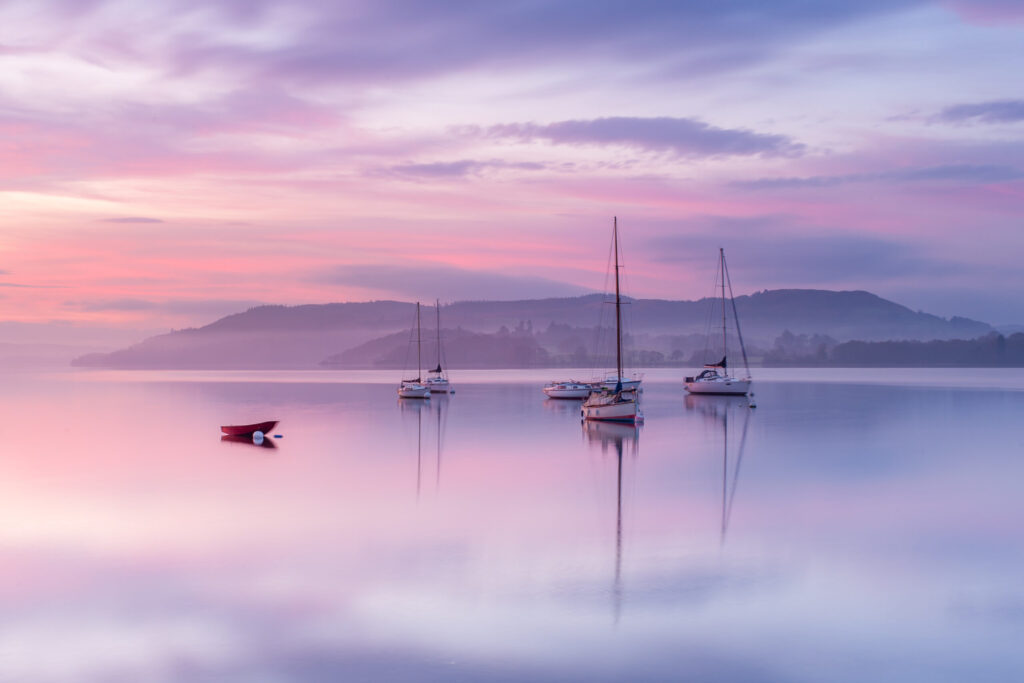Moored sailing boats on a calm glassy windermere lake reflecting a bright pink and purple sunrise