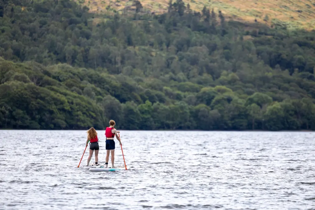 Two young adults paddle boarding on a lake with a woodland backdrop