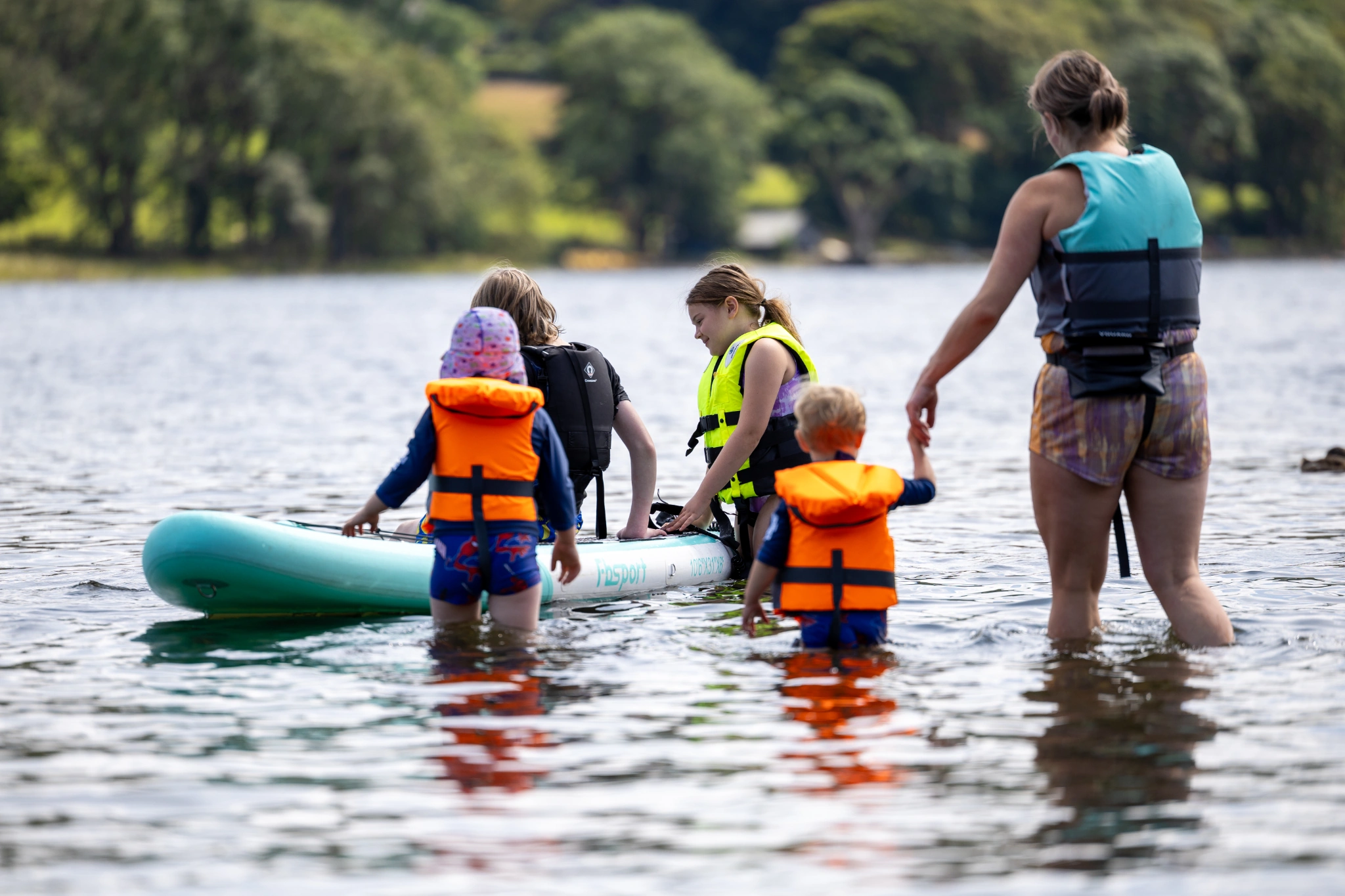 Young children stood in the shallow lake water guided by two adults walking towards a paddle board, all wearing buoyancy aids