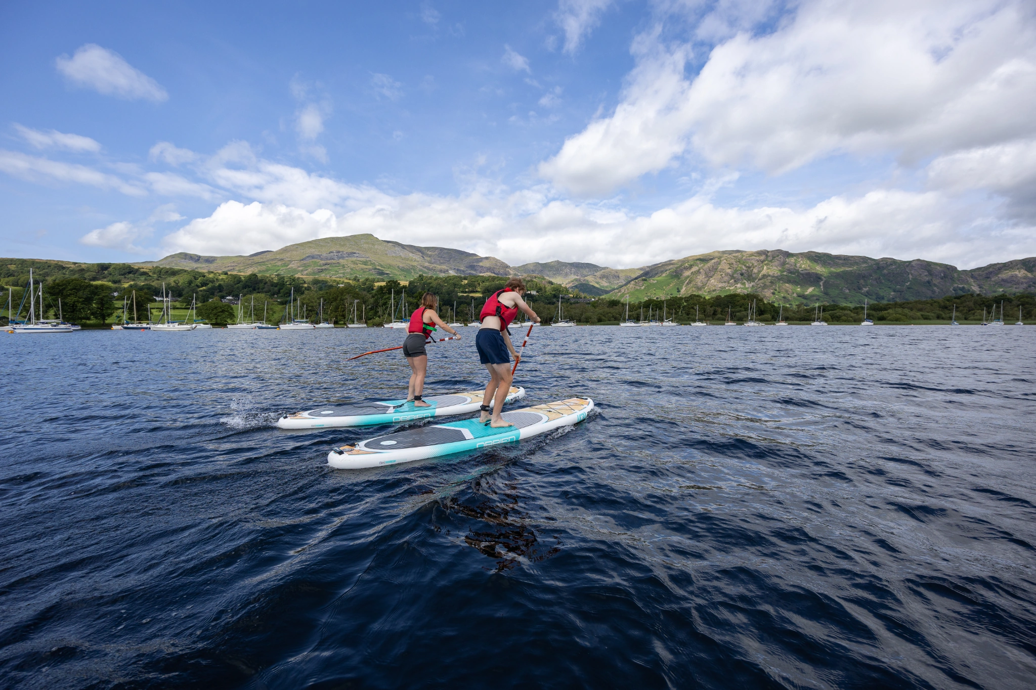 two young adults wearing summer active clothes and red buoyancy aids on turquoise paddle boards in the lake, with a green woodland and fell backrop