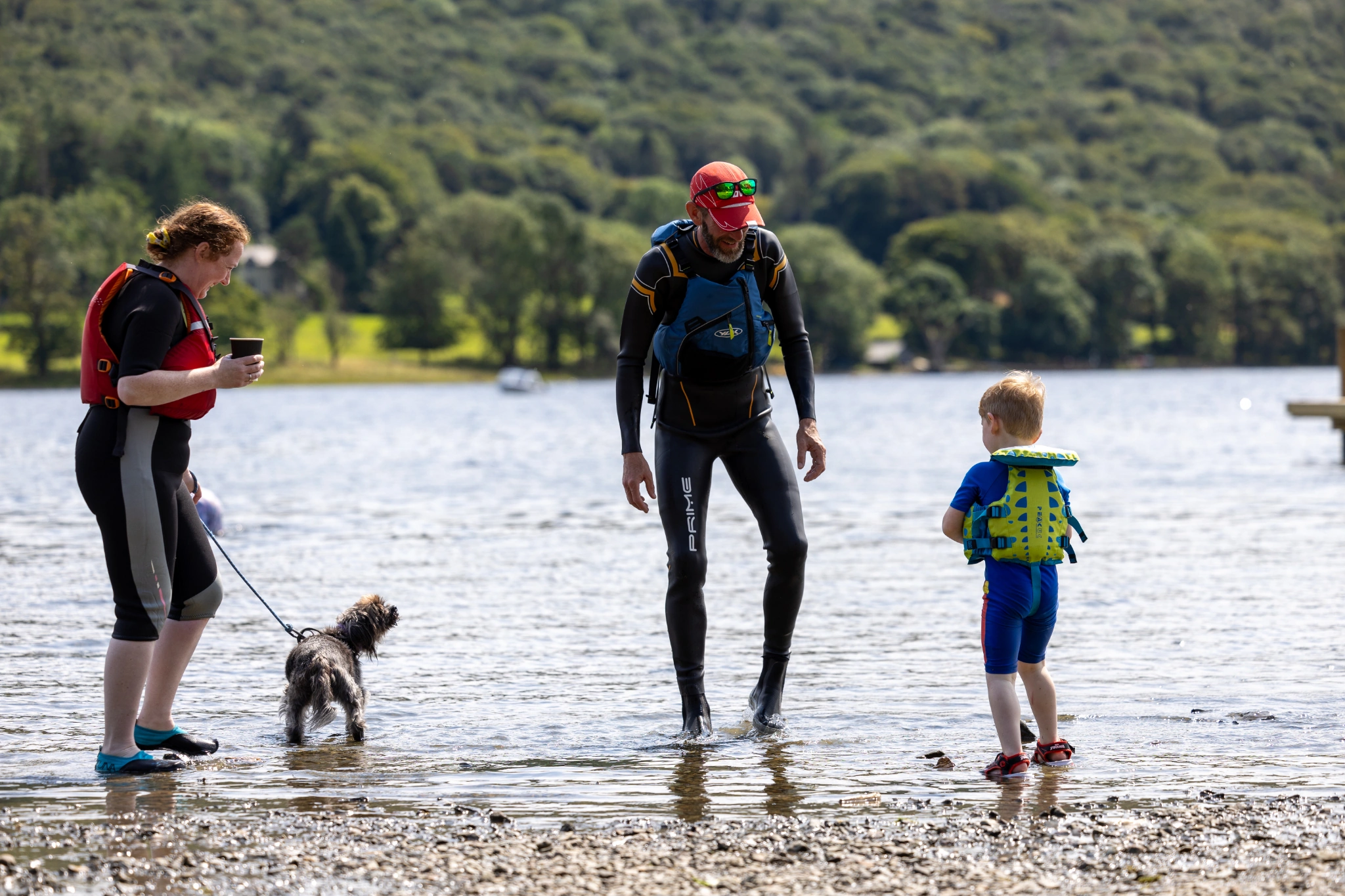 Adult woman with a small dog on a lead, adult male and young boy in wetsuits and buoyancy aids stood in the shallow lake shore water. green woodland backdrop