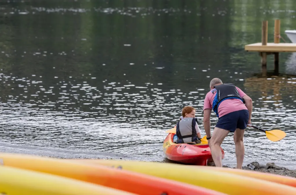 Father pushing teen in kayak out onto the lake, both wearing summer clothes and buoyancy aids, with stored orange and yellow kayaks in the foreground