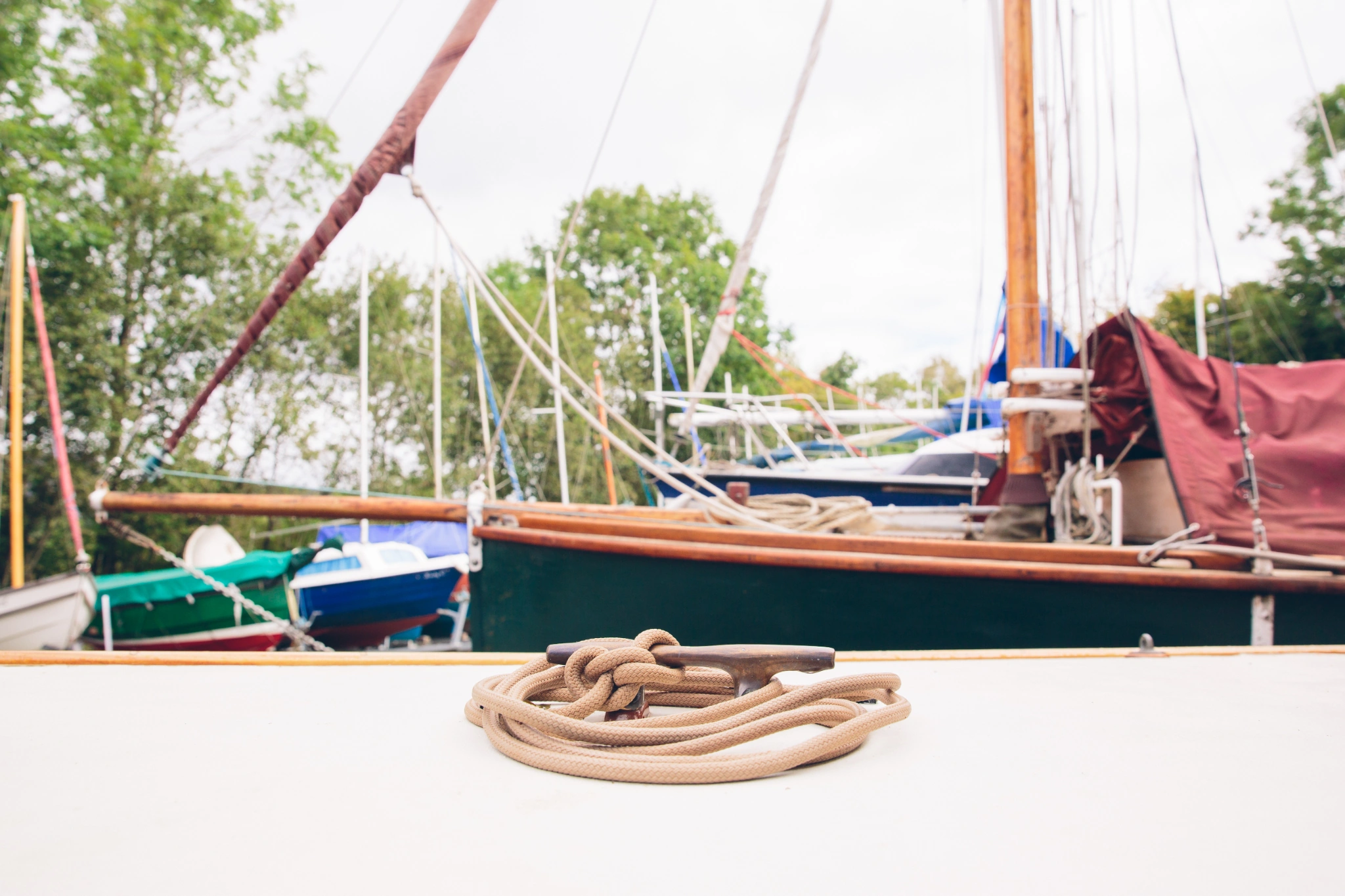 Stored colourful sailing boats with a boat deck and coiled rope in the foreground and green trees in the background